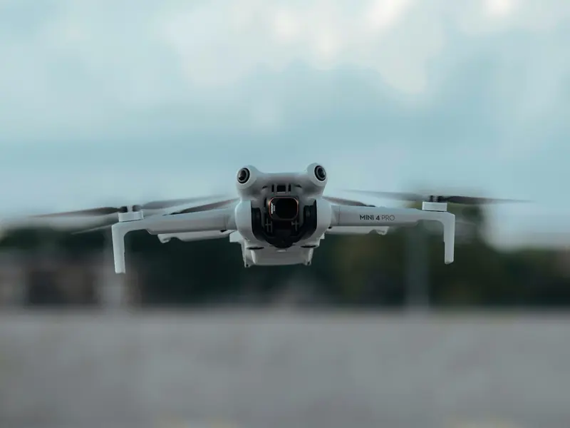 Un drone DJI Mini 4 Pro gris clair en vol, vu de face, sous un ciel nuageux