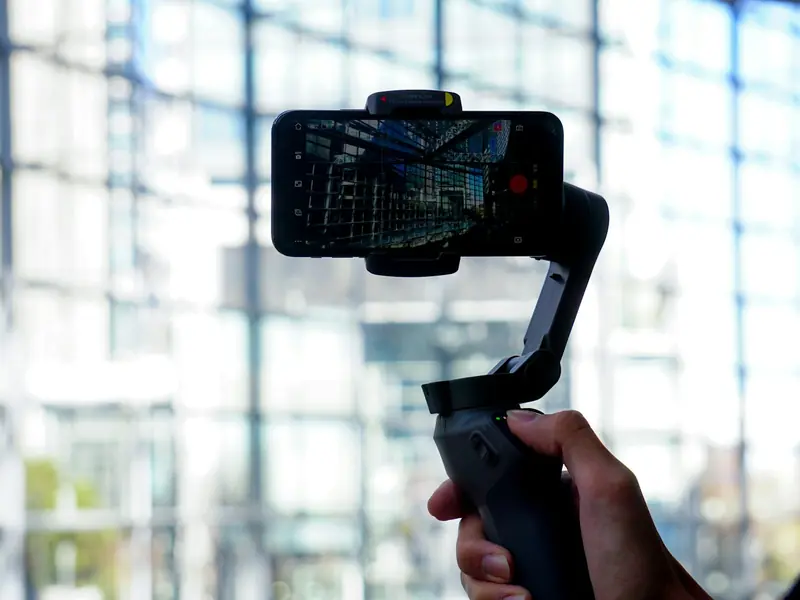 Un stabilisateur de téléphone DJI Osmo Mobile tenu à la main, un smartphone en train de filmer fixé dessus