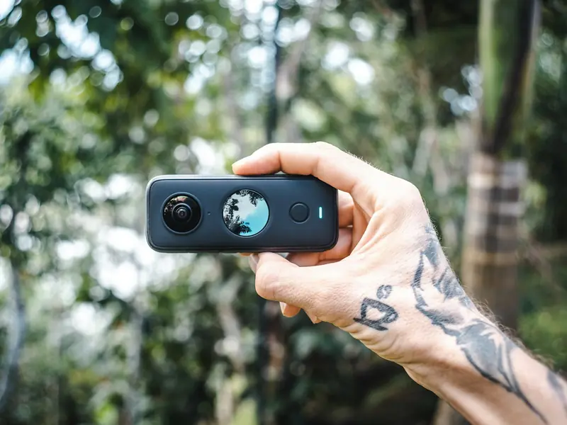 Une caméra 360° Insta360 noire à deux objectifs tenue à la main devant une végétation tropicale