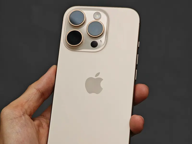 Un iPhone 16 Pro couleur titane sable, vu de dos avec ses trois objectifs, tenu dans une main